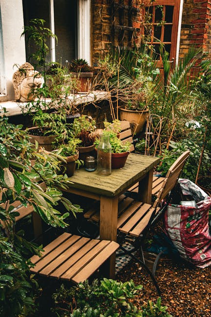 A small urban garden space featuring a weathered wooden outdoor dining table surrounded by matching wooden chairs, set on a compact gravel surface. The table is decorated with various potted plants, i