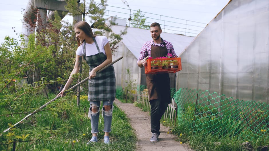 A young woman and a man are engaged in garden maintenance along a narrow pathway, surrounded by lush greenery and plants. The woman, wearing a white t-shirt, ripped jeans, and a green and black checke