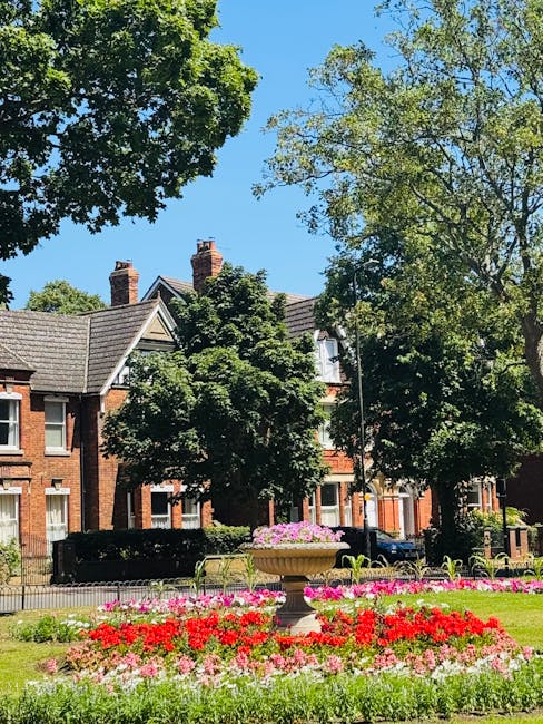 A well-maintained urban garden scene featuring a vibrant flower bed in the foreground with red, pink, and white blooms, bordered by lush green grass. Behind the flower bed, there is a decorative stone
